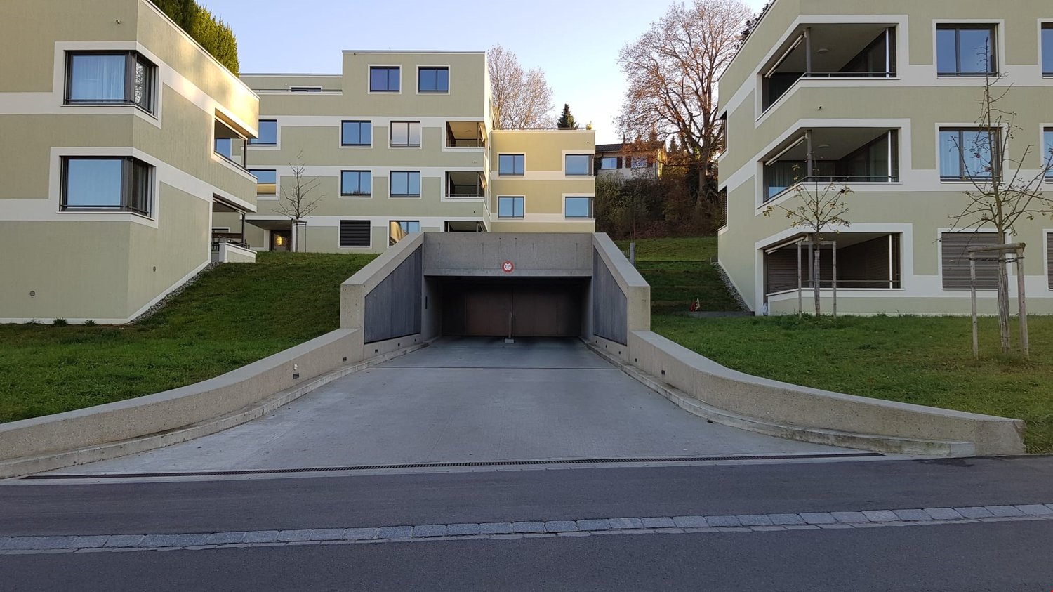 Multi-story apartment building with a garage entrance in the foreground. The building has a modern, minimalist design with a mix of yellow and gray colors. There are balconies and large windows visible on the facade. The surrounding area appears to be a r