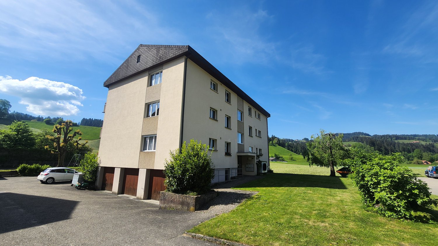 Multi-story building, beige walls, multiple windows, car parked in front, greenery surrounding, hills in the background