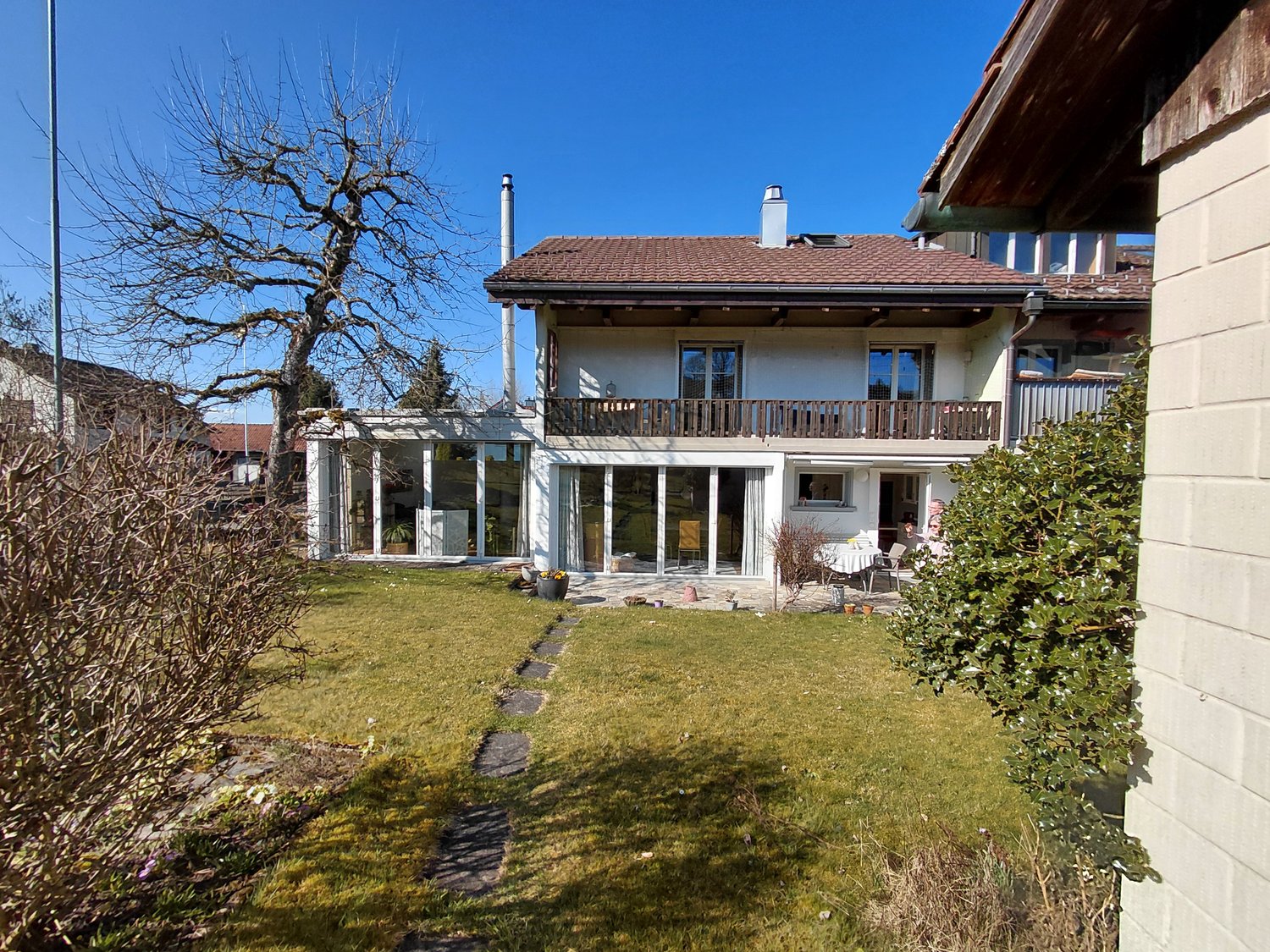 2-story house, brown roof, large glass windows, balcony, garden, potted plants, pathway, grassy lawn, trees, nearby houses