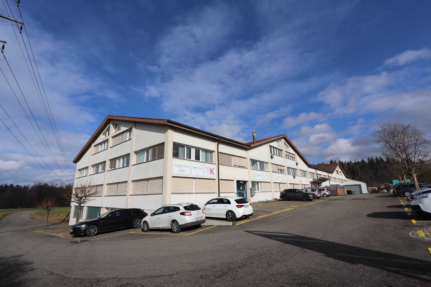 Two-story white building, signboard, several parked cars, trees in front, surrounding landscape