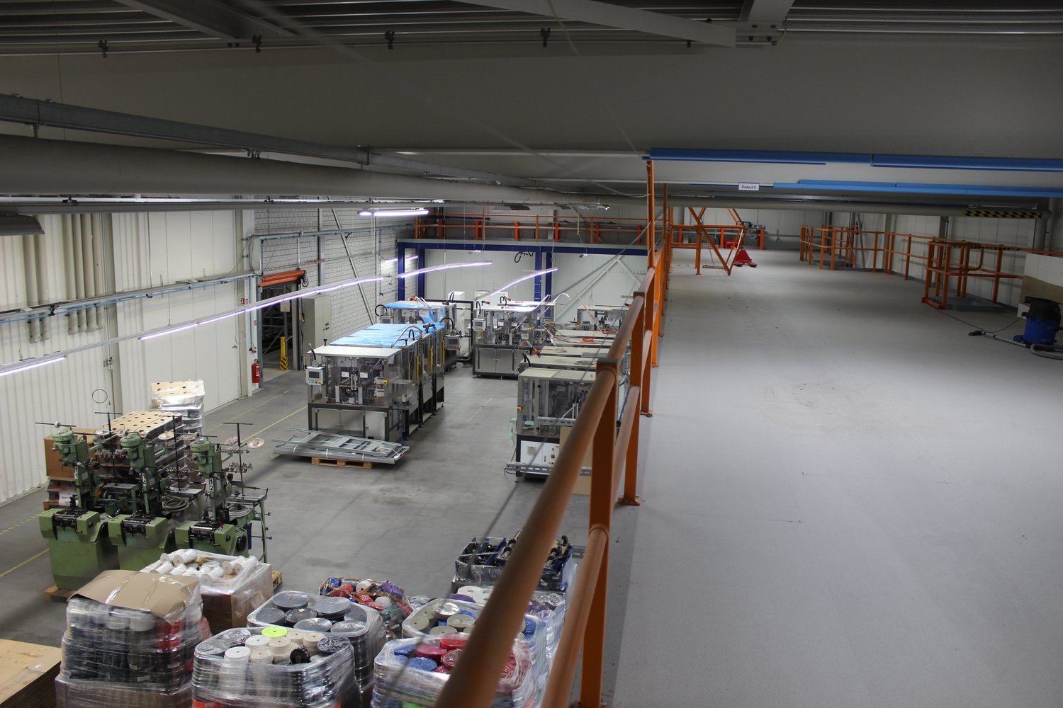 The image shows a large industrial facility or warehouse. The space is filled with various equipment, machinery, and storage racks. There are several workstations and production lines visible. The ceiling is high and the lighting is industrial. The overal