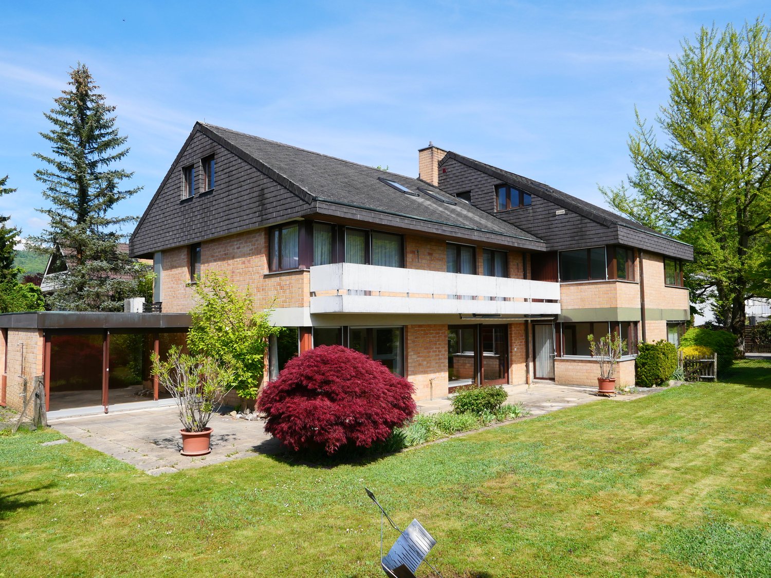 2-story house with a slanted roof, brick and wood exterior, large windows, balconies, and a well-maintained garden with a red Japanese maple tree in the front yard. There appears to be a parking area or driveway in front of the house.