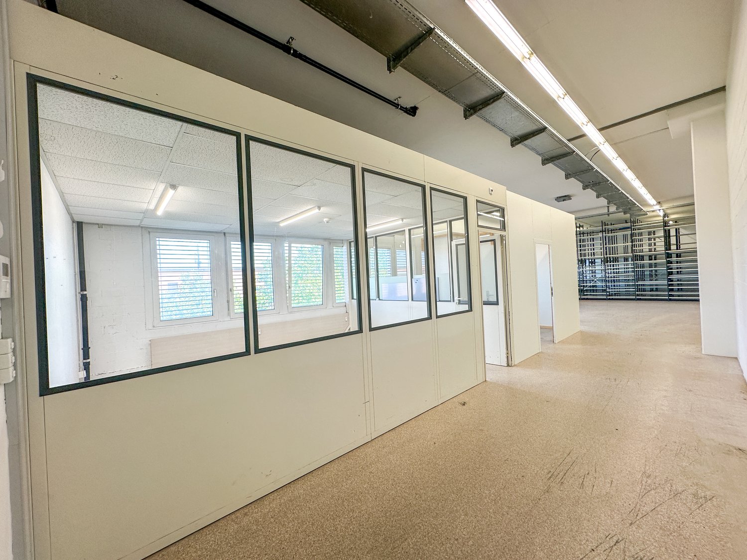 The image shows a large open office space with white walls, windows, and a concrete floor. The space appears to be unfinished, with exposed ceiling beams and lighting fixtures. The room has multiple glass-paneled walls, creating separate sections or offic