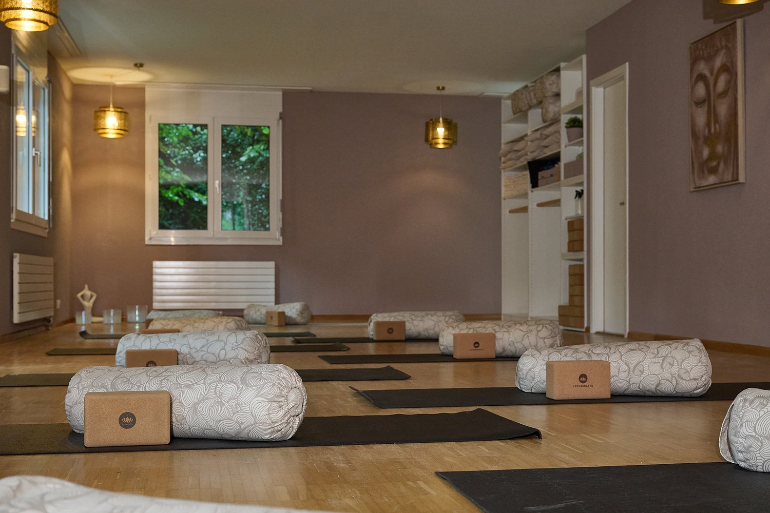 A yoga studio with yoga mats, bolsters, pillows, and lamps on the ceiling. The floor is wooden, and the walls are painted in a muted purple. There is a shelf with towels, a potted plant, and a framed artwork.