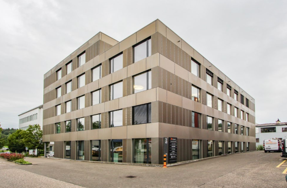 A multi-story office building with a modern, angular design. The exterior is made of beige and brown panels, with large windows throughout. There is a parking area visible in the foreground of the image.