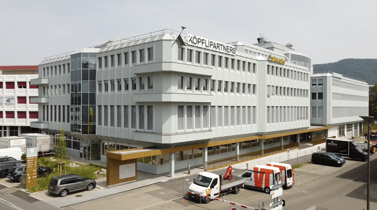 white exterior, multiple windows, 'Koepfli Partners' sign, cars parked in front, adjacent buildings, mountains in the background