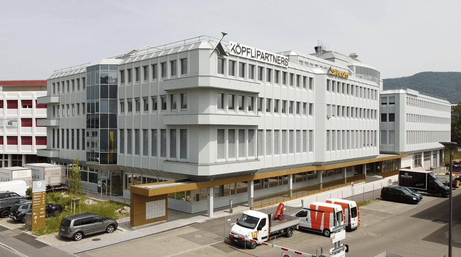 multi-story office building, white exterior, glass windows, red sign reading 'KOPFLI PARTNERS', multiple parked vehicles in the front