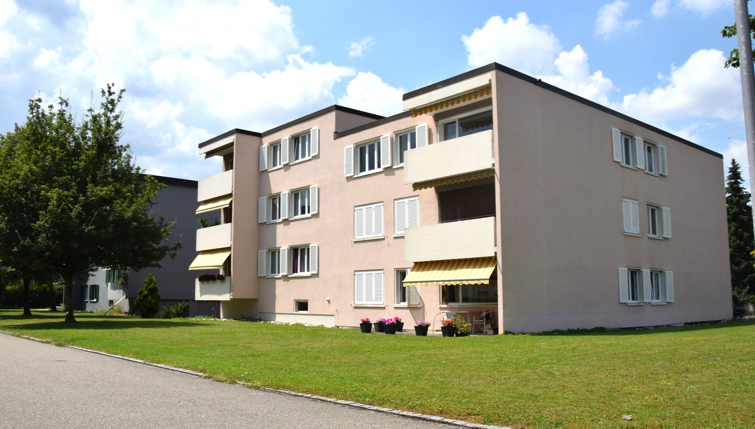 3 story apartment building, beige exterior, white windows, yellow awnings, balconies, front yard with grass and trees