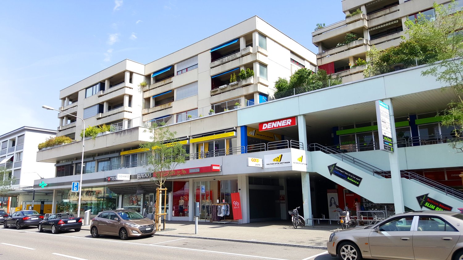 Multi-story commercial and residential building with balconies, parking spaces, and various retail stores on the ground floor including Denner supermarket.