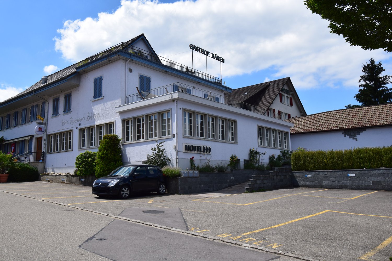 Hotel with white walls, black roof, balcony, several windows, sign above the building, parking lot