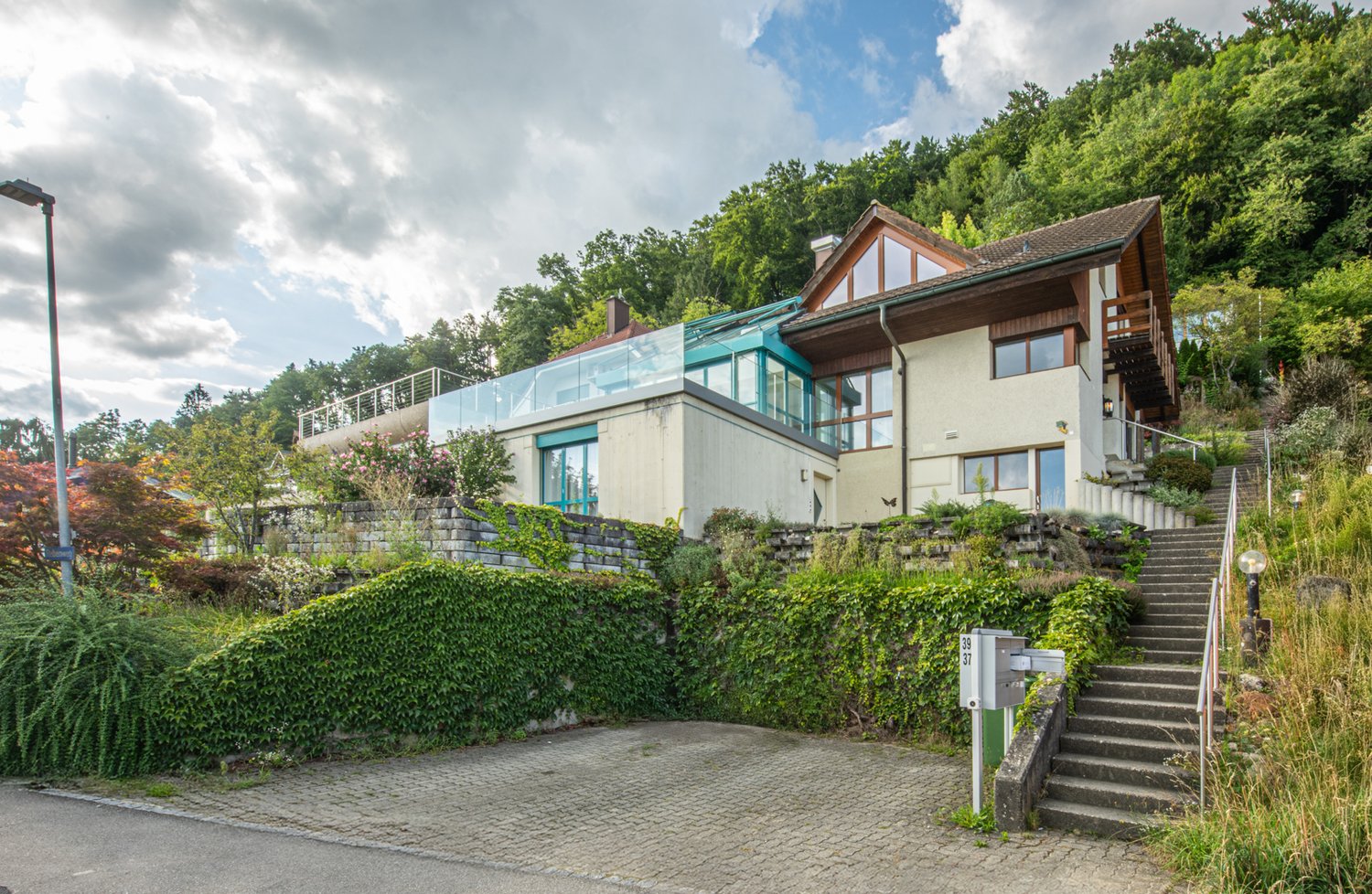 House on slope, glass balcony, terrace, brick ground floor, driveway, front and side gardens