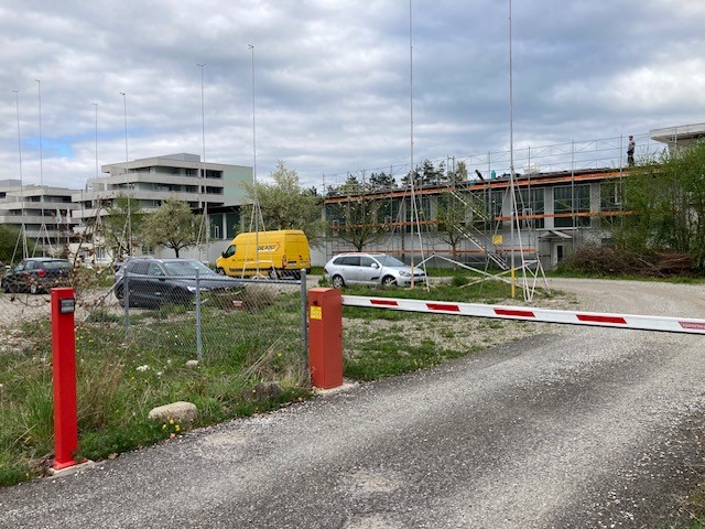 Empty parking lot, 4 cars parked, red barrier gate, fence with posts, buildings in background, cloudy sky, grass and shrubs on ground.