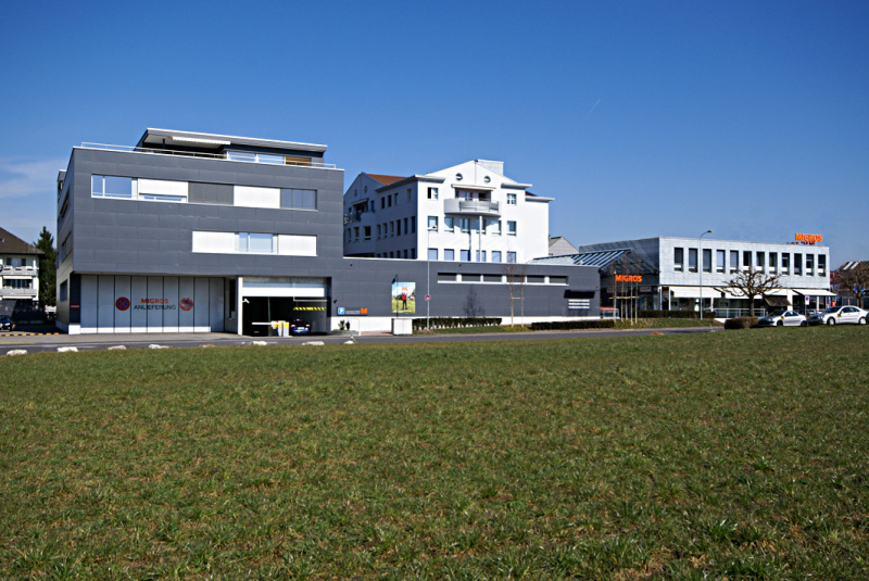 Commercial building with sign 'Migros', grey exterior, multiple windows, open garage door, cars parked in front, spacious lawn area