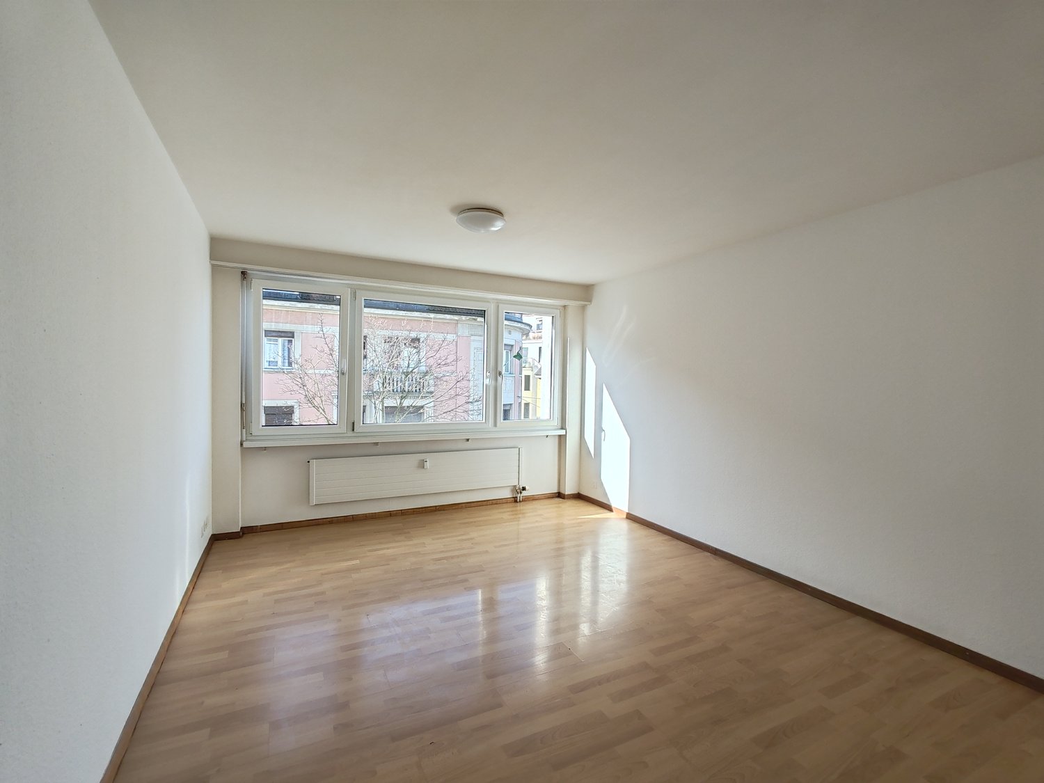 Empty room with white walls, wooden floors, and large windows with view of the outside.
