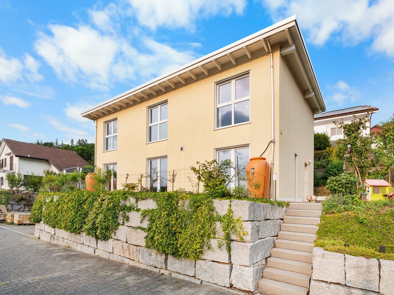 Two-story house, yellow exterior, white windows, stone wall, large staircase, fenced area, green plants, orange vase.