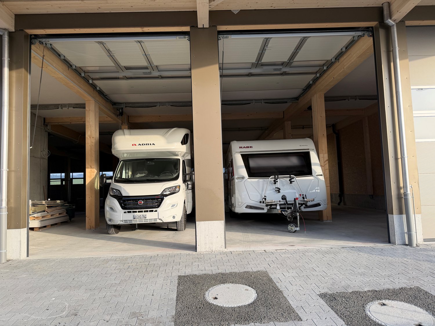 Two vehicles parked in a garage, one is a camper van and the other is a trailer.