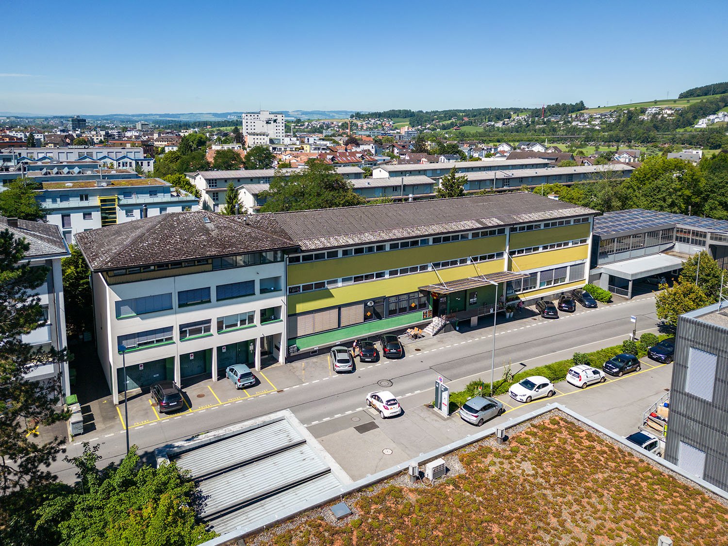 Large office building with yellow and green exteriors, numerous windows, parking lot with cars parked, solar panels on the roof, surrounded by greenery and other buildings