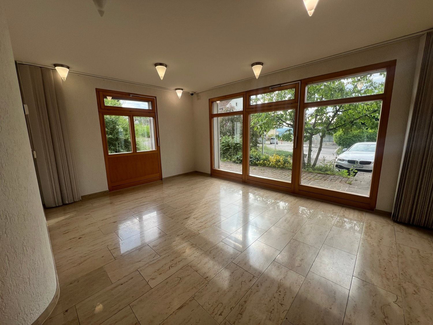 Spacious living room with large windows, hardwood floors, and recessed lighting. The room has a bright and airy feel with views of the surrounding greenery outside.