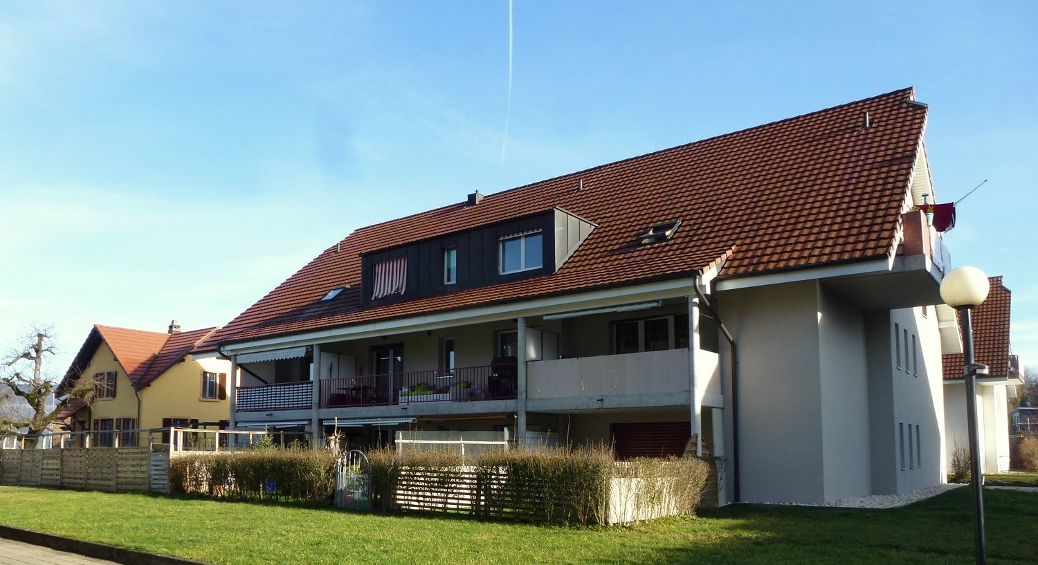 3 story residential building, brown roof, balconies on 2nd and 3rd floor, fence, grass