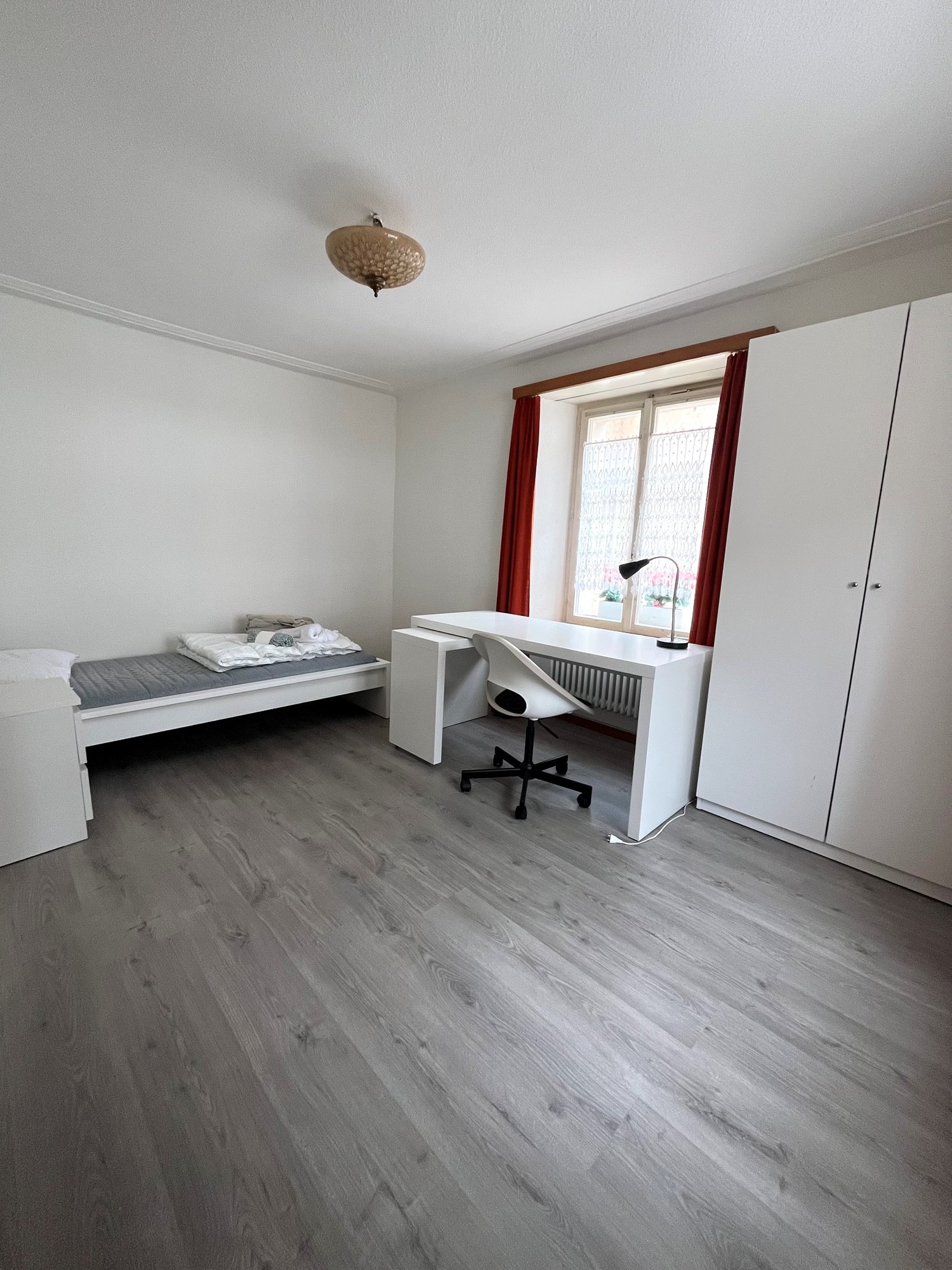 room features a single bed, desk with a chair, a window with red curtains, and a lamp. White walls and wooden floors.