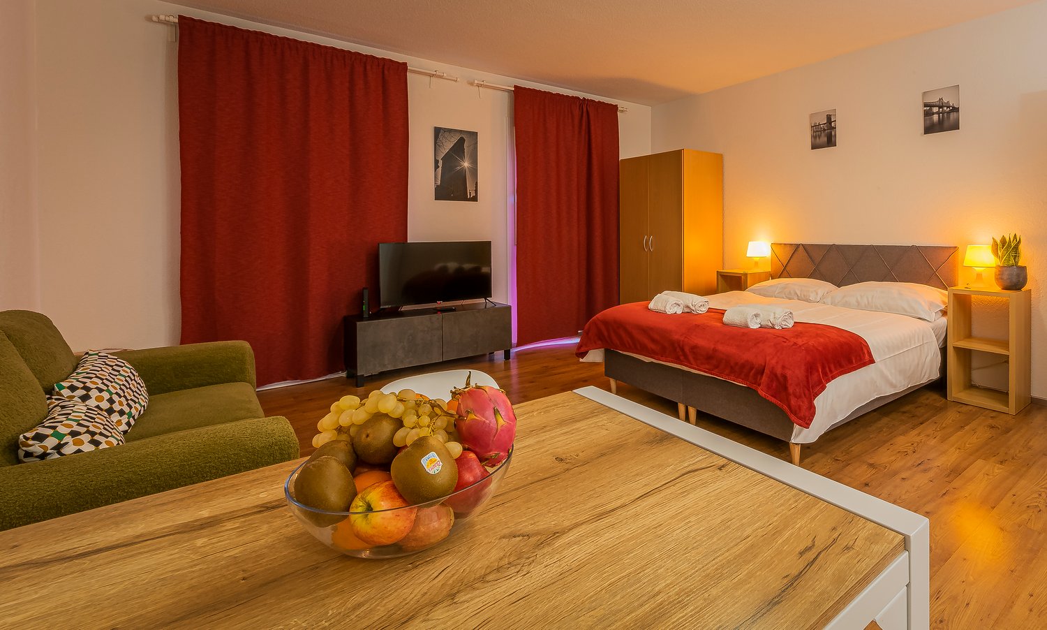 Bedroom with wooden floor, a bed, a TV, a green couch, a table with fruits, and nightstands with lamps and a potted plant