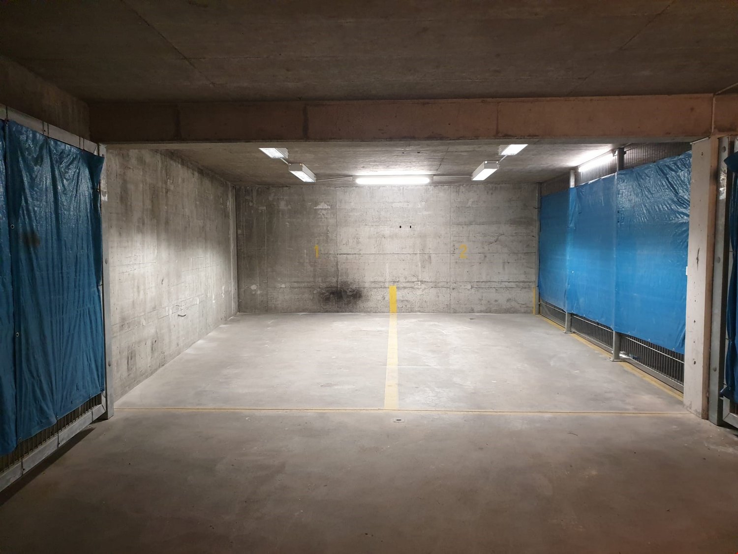concrete walls, gray floor, yellow marking lines, blue plastic barriers, ceiling lights
