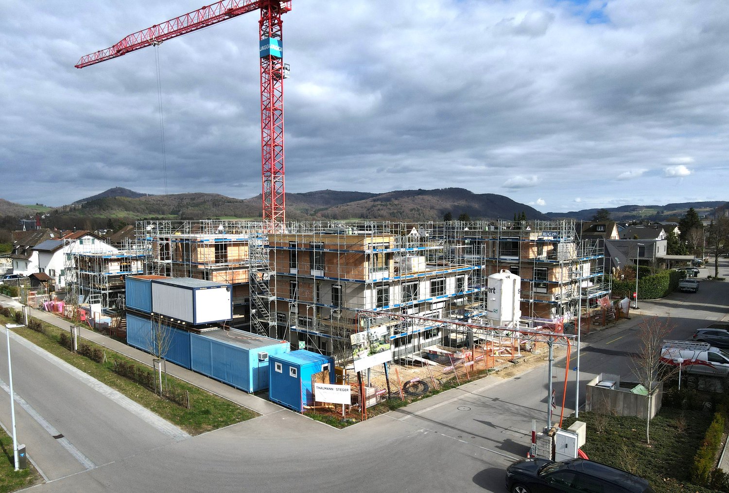 Aerial view of a construction site with multiple buildings under construction, a large red crane, scaffolding, and a road with parked cars.
