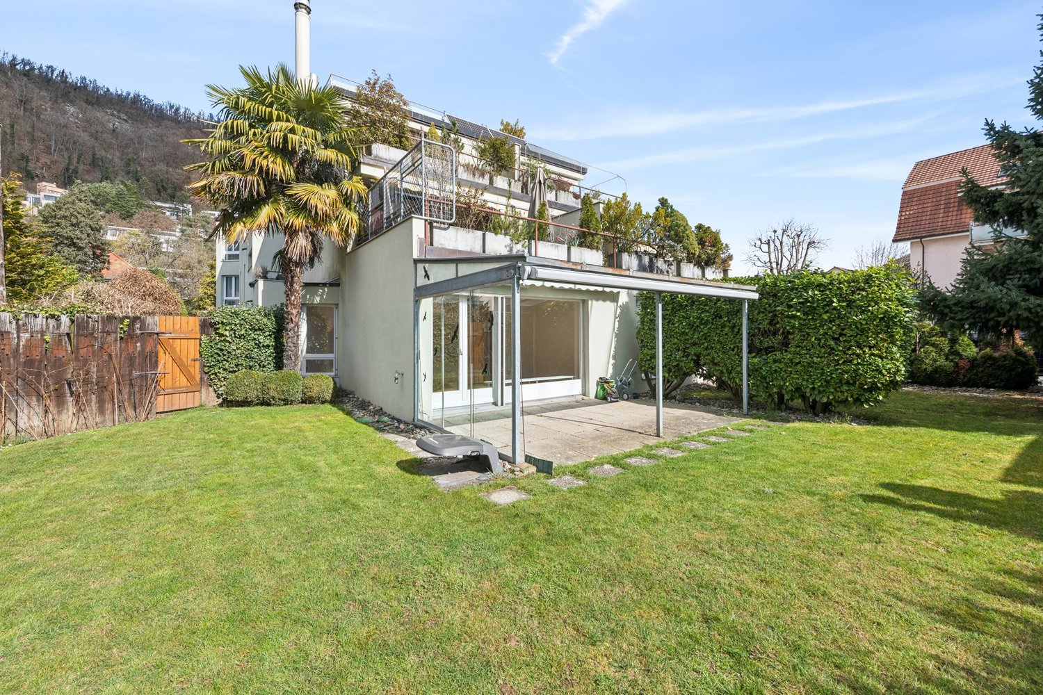 Modern house with a terrace, glass doors, and a carport, surrounded by a well-maintained garden with a lawn, bushes, and a palm tree.