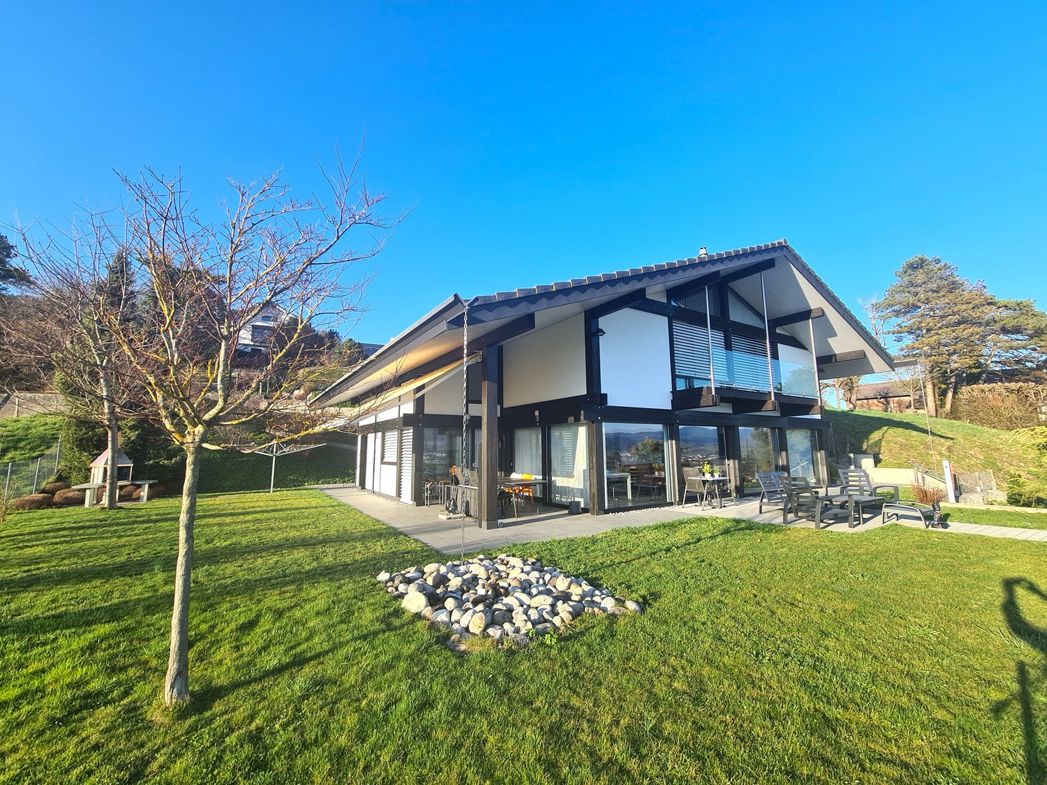 House with modern design, large windows, terrace with seating area, lush lawn, patio with fireplace, surrounded by greenery.