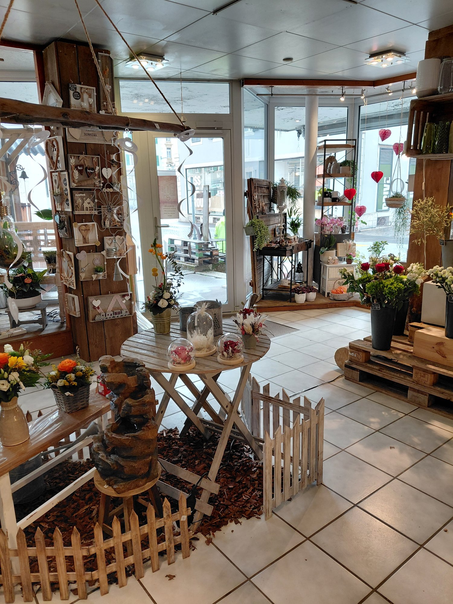 Room filled with decorative items and floral arrangements, white tiled flooring, wooden tables and benches, natural light from windows