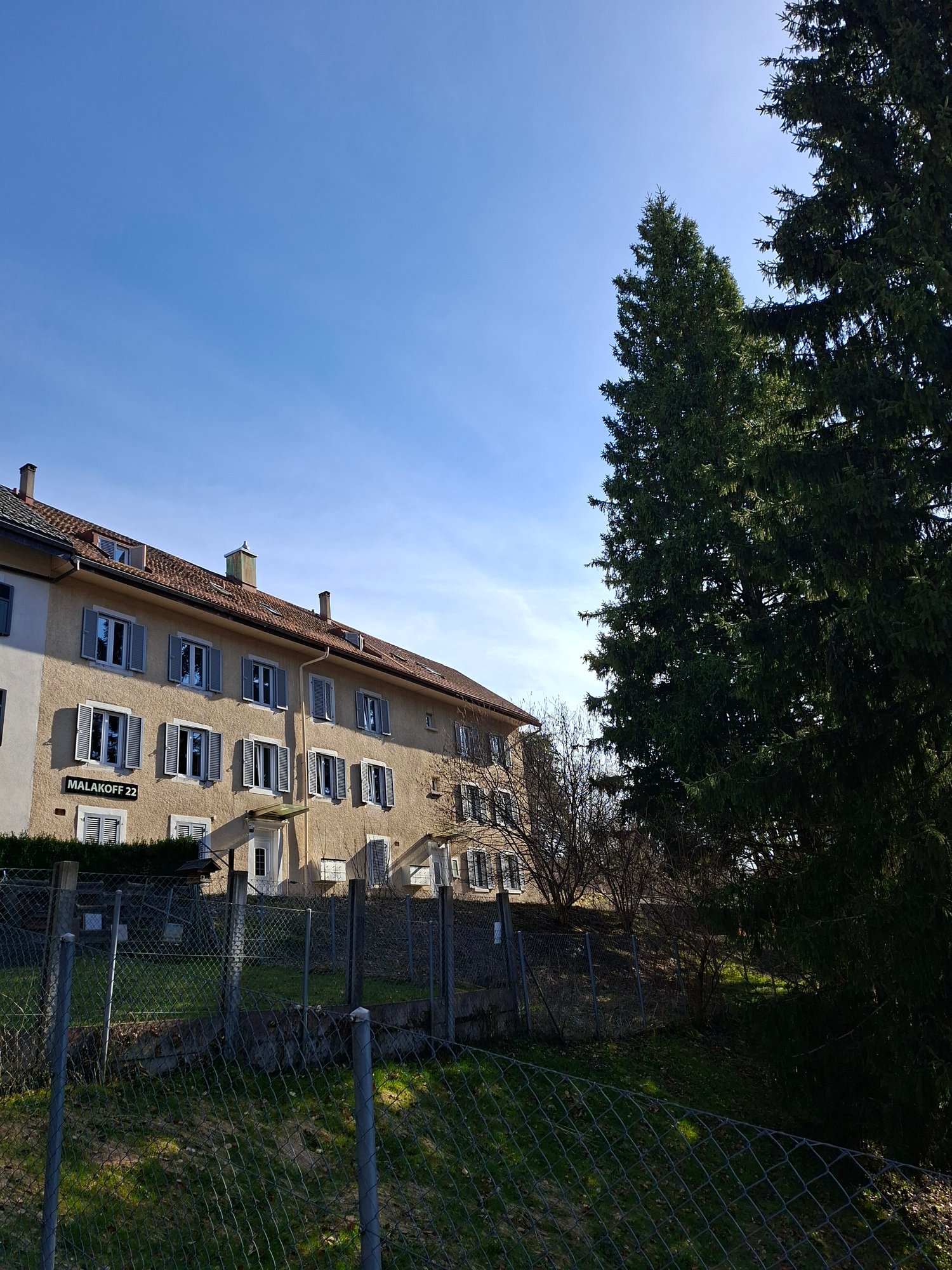 3 story building, brown roof, multiple windows, sign reading 'MALAKOFF 22', fenced yard, trees