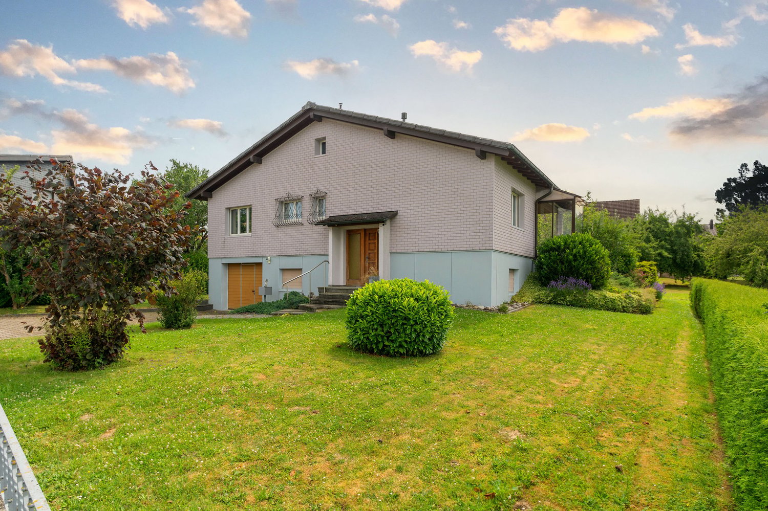 2-story house with white brick exterior, large windows, wooden front door, attached garage, surrounded by a well-maintained lawn with shrubs and trees