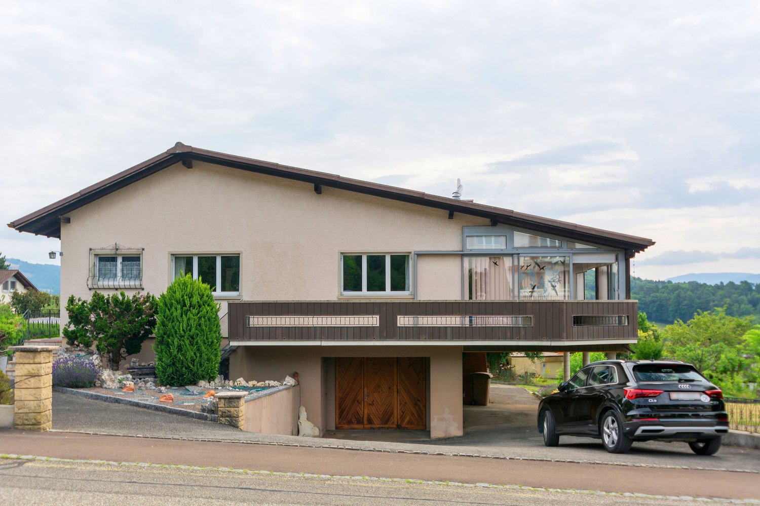 2-story house with stucco exterior, brown roof, large windows, wooden garage door, balcony, and a parked car in the driveway. The house is surrounded by landscaping with shrubs and trees.
