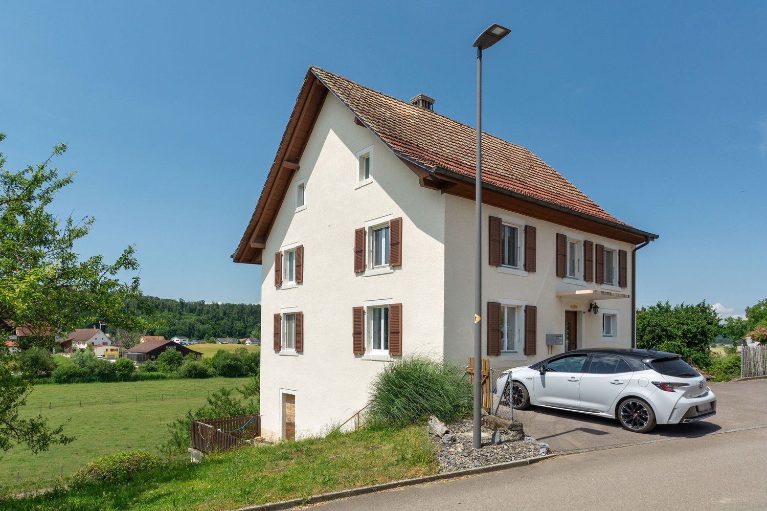 2-story white house with red tile roof, brown shutters, and a parked car in the driveway. The house is surrounded by green trees and grass.