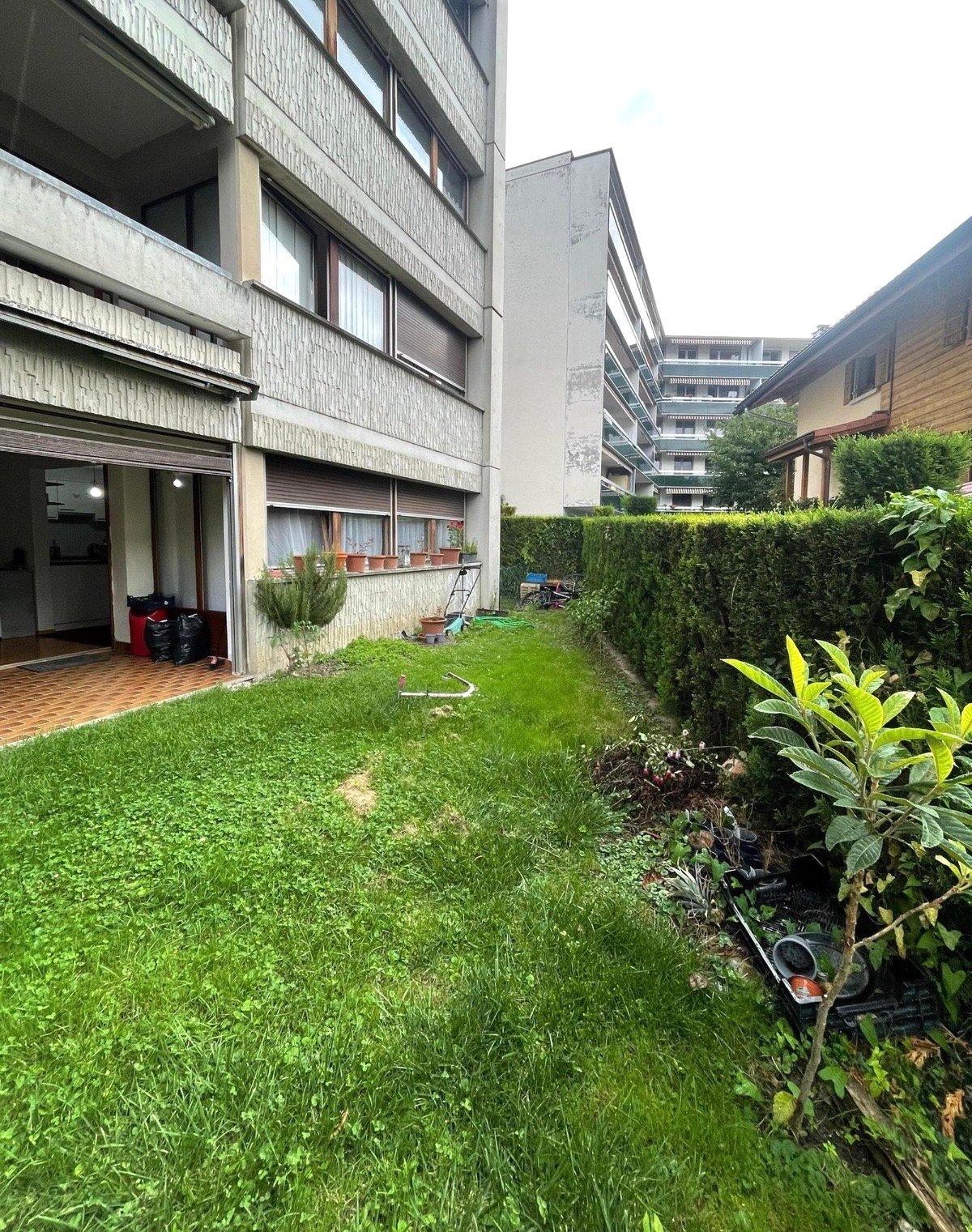 Building with tiled entrance, open garage, small garden area, balcony with potted plants, hedge, grass lawn, trash bins, and a tool cart.