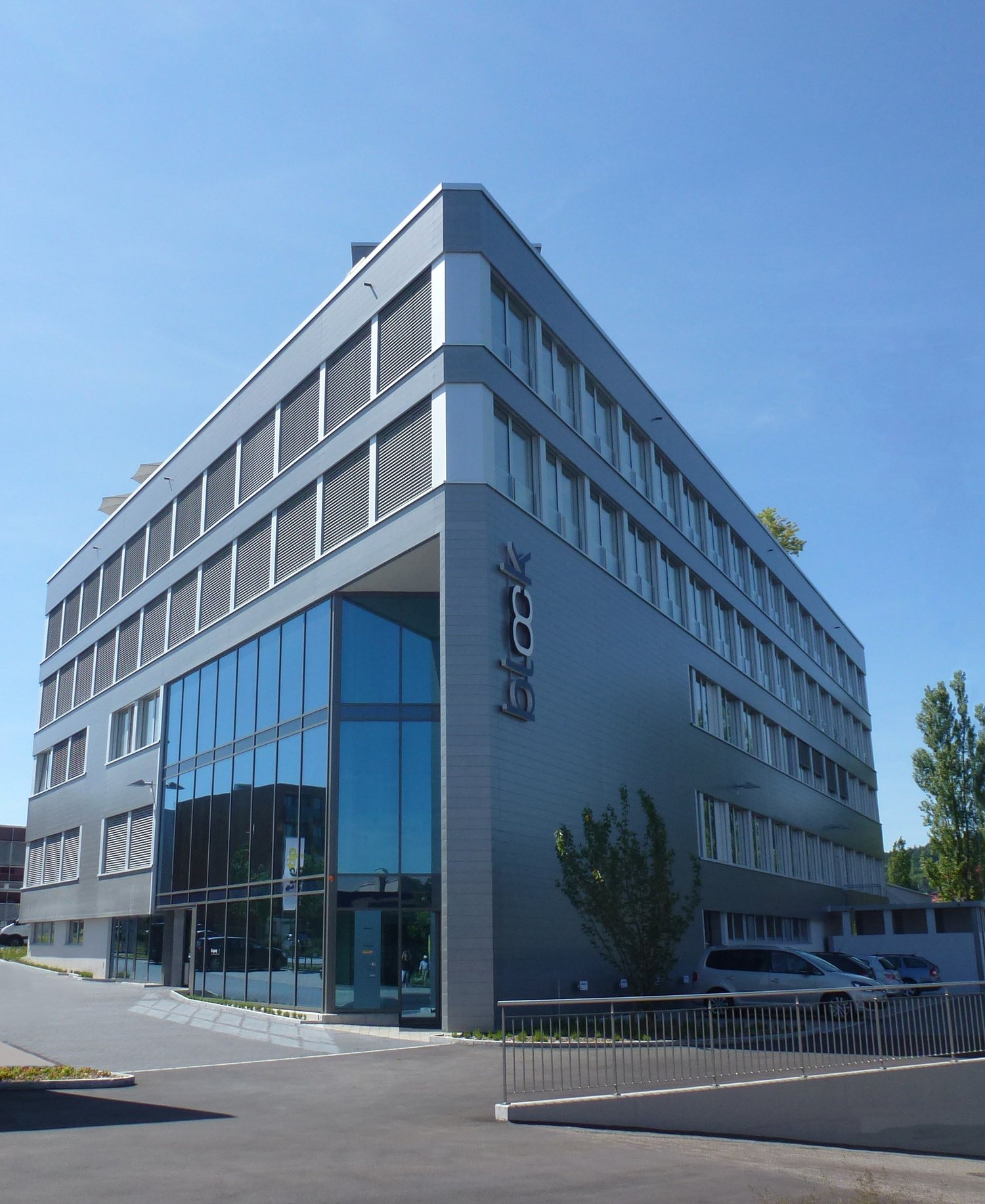 Modern office building, grey panels, large windows, multiple cars parked outside