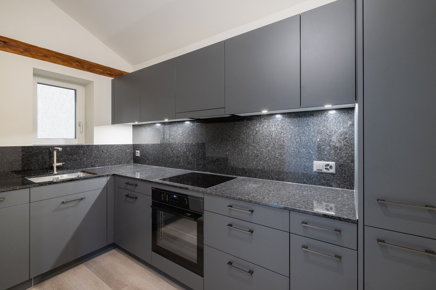 Modern kitchen with grey cabinets, granite countertops, built-in oven, induction cooktop, and modern fixtures.