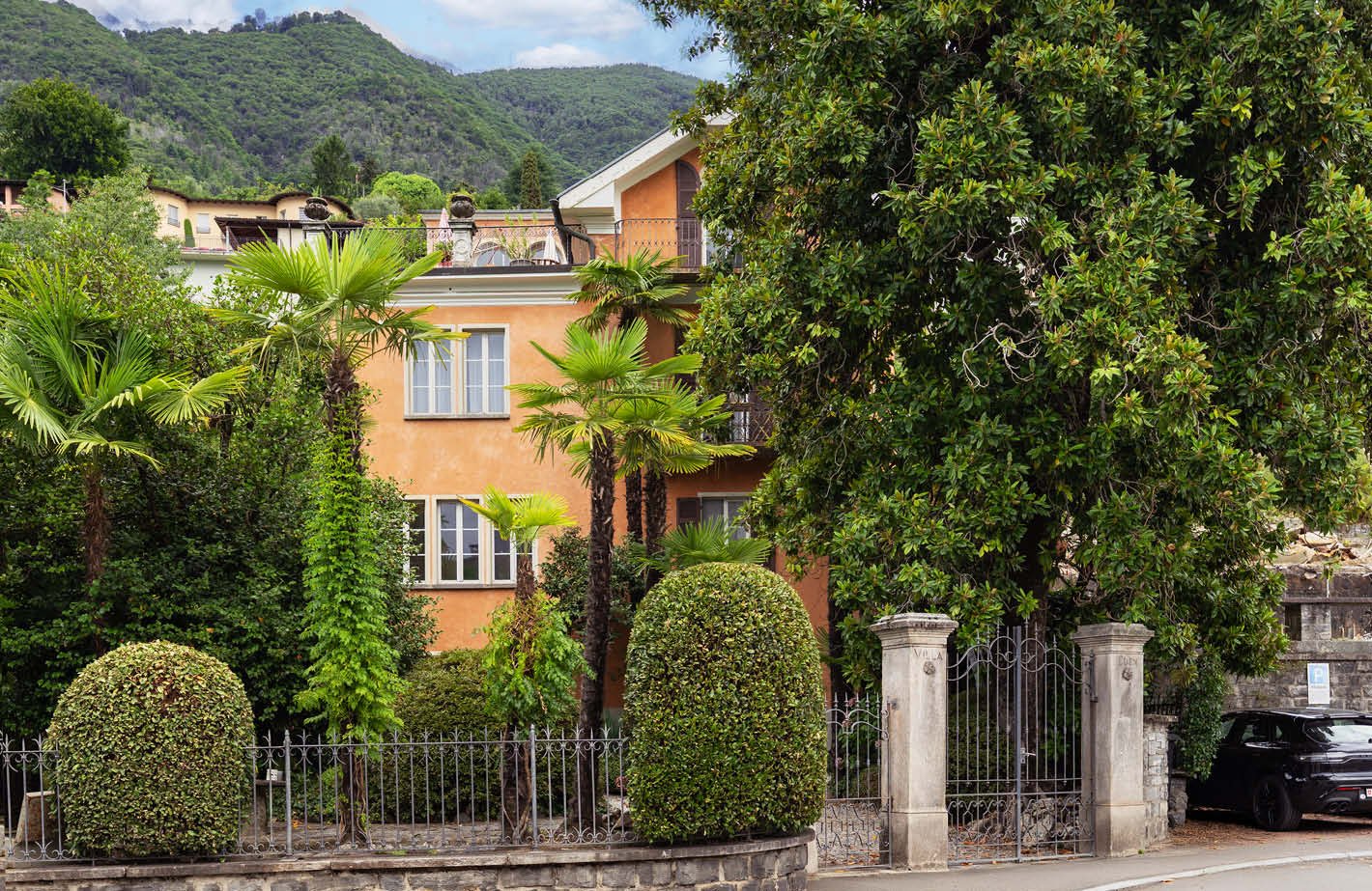 A detached house with a balcony on the second floor, white walls, and brown shutters. Palm trees and hedges adorn the front of the house. In front of the house, there is a paved sidewalk with iron railings and a parking area with a black car.