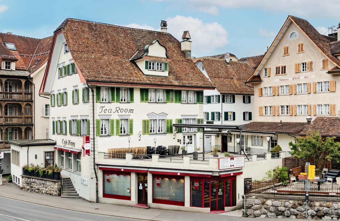 A multi-story building with a traditional European architectural style, featuring red-tiled roofs, green shuttered windows, and a balcony. The building houses a tea room on the ground floor, with outdoor seating. The surrounding buildings have a similar s