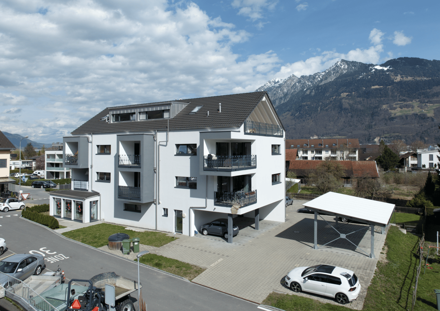 4-story apartment building with white exterior, slanted roof, balconies, and parking spaces in the front