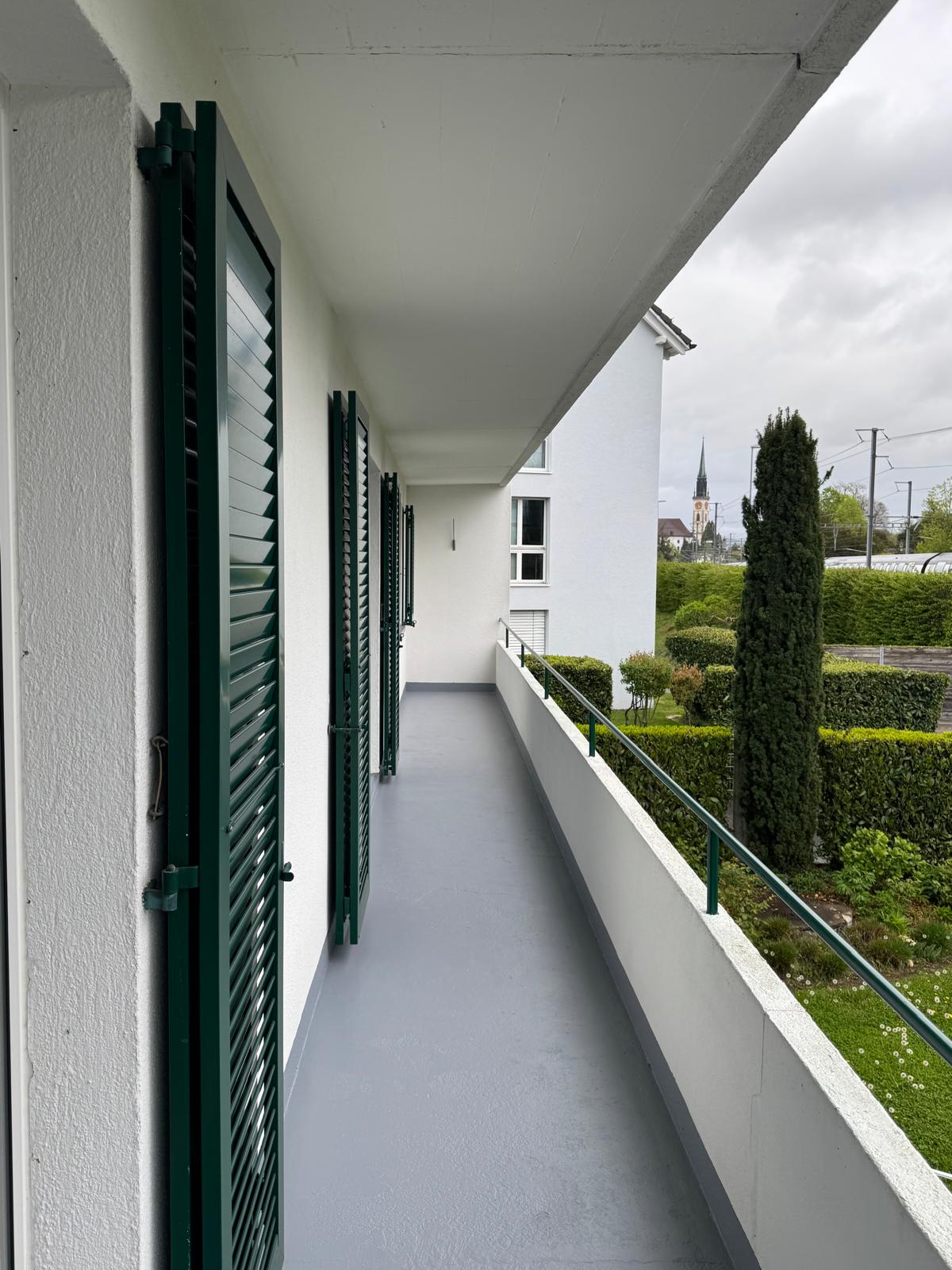 balcony with green shutters, gray floor, white walls, green railing, view of a garden