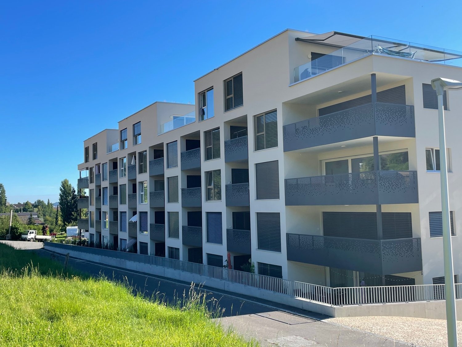 Modern apartment building, multiple floors, balconies, glass railings, white exterior, green grass, street view