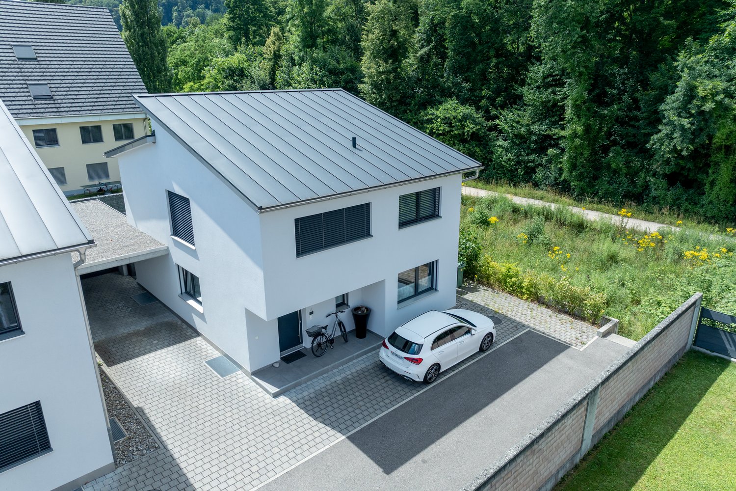 Modern 2-story house, white exterior, gray metal roof, driveway, bicycle parked in front, white car parked in driveway, fenced garden, trees and plants in the background