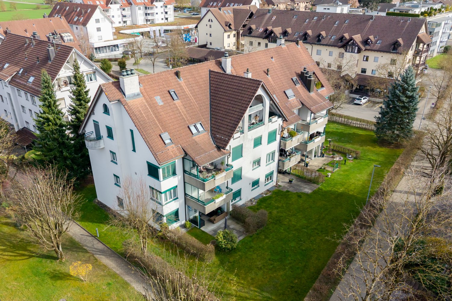 Aerial view of a multi-story residential building with brown roofs, white walls, and green window frames. Balconies are visible on the upper floors. The building is surrounded by a well-maintained green lawn and several trees. A car is parked in the parki