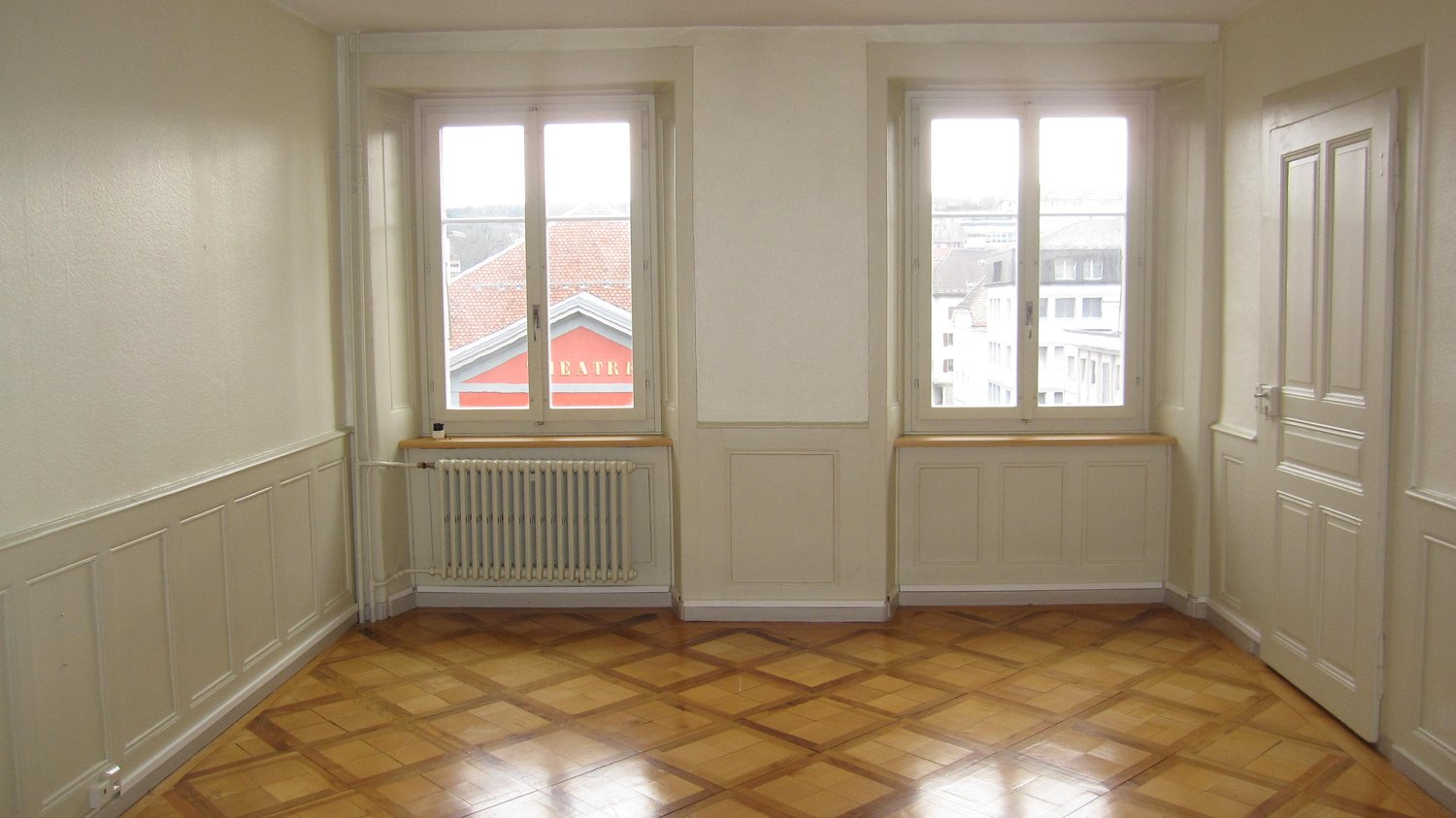 Empty room with wooden parquet flooring, white walls, two windows, two white radiators, door at the end
