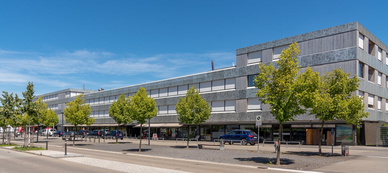 Modern building with multiple floors, gray exterior, large windows, parking lot, trees, cars parked in front