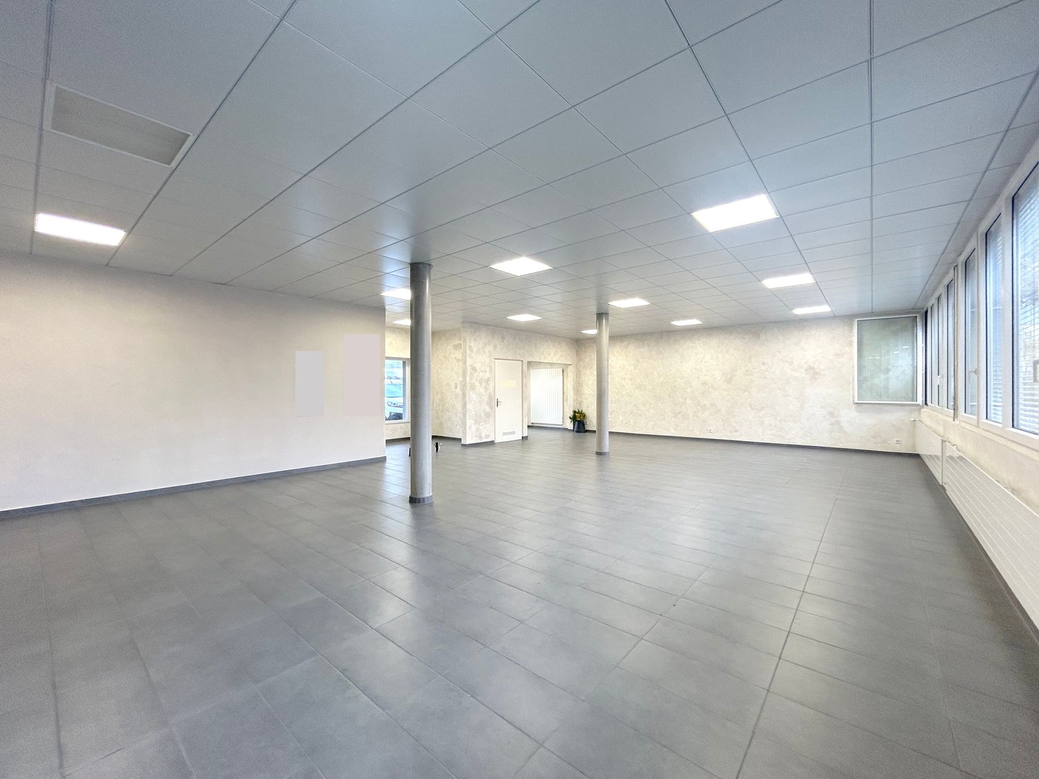 Empty room with grey tiles, white walls, square ceiling lights, and windows with blinds.
