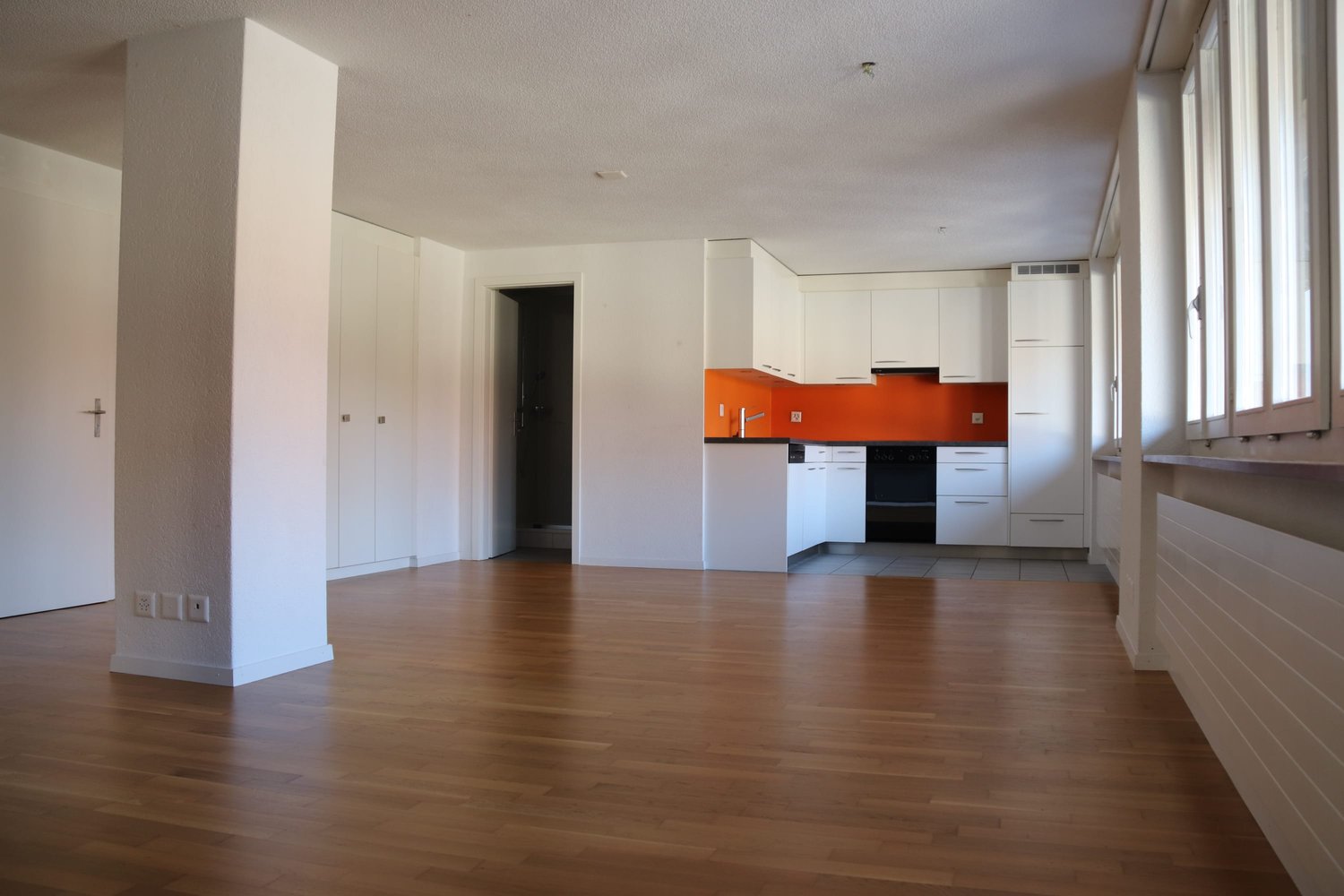 empty room, wooden floor, white walls, white cabinets, orange wall in kitchen area, large windows