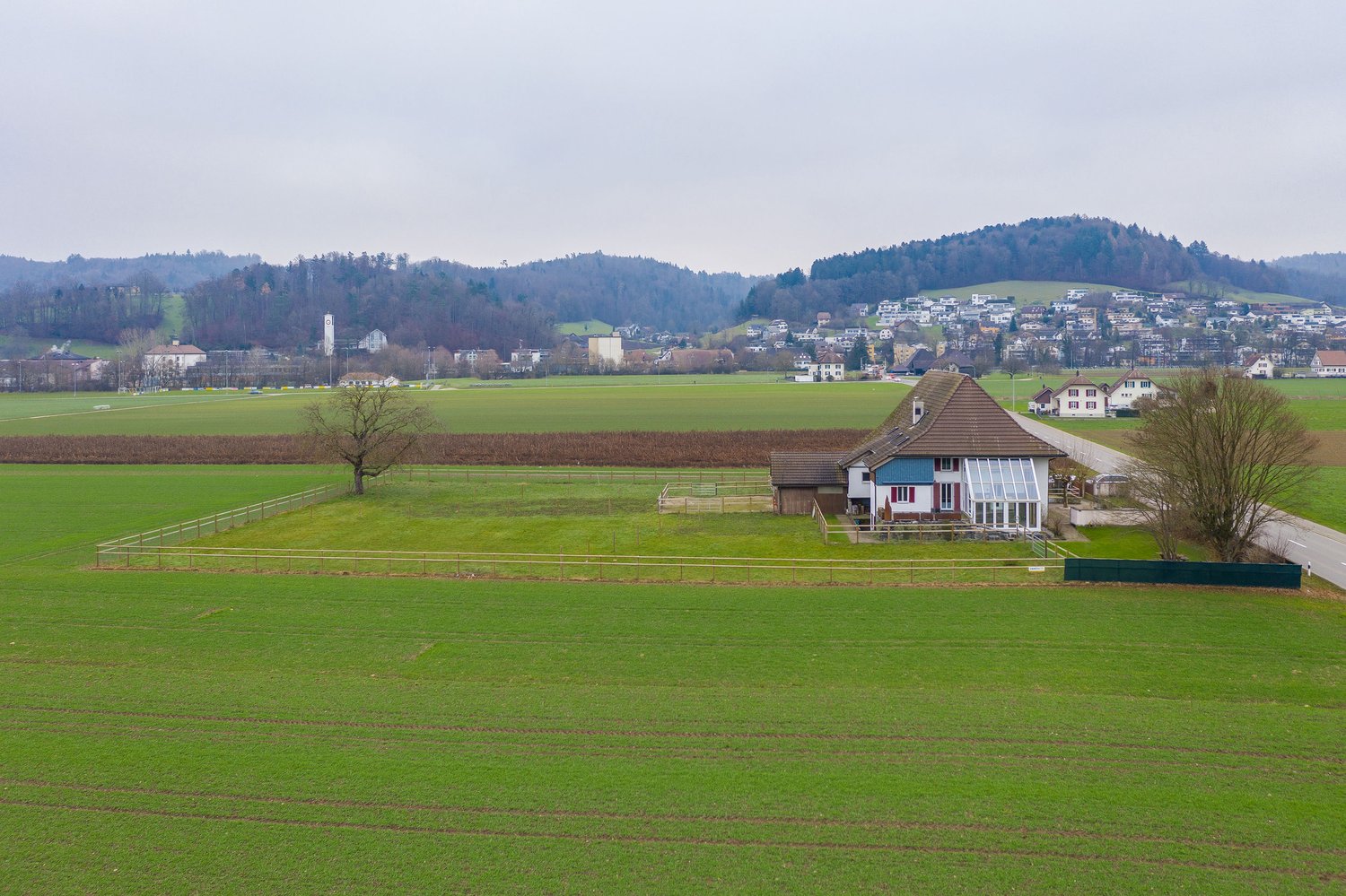 The image shows a rural countryside landscape with rolling green hills, a small village in the distance, and a farmhouse with a balcony or terrace in the foreground. The scene has a peaceful, pastoral feel with fields, fences, and a few trees dotting the