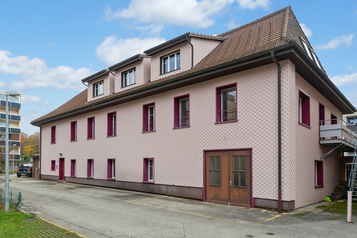 pink facade, brown roof, multiple windows, ground floor entrance, parking area, balcony
